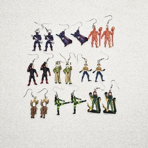 May include: A collection of handmade earrings featuring miniature figurines. The earrings showcase a variety of characters in different colors, including soldiers, superheroes, and other fantasy figures. Each earring has a silver hook.