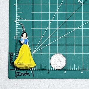 May include: A pair of silver earrings featuring a plastic figurine of Snow White in a yellow dress with a blue bodice. The earrings are approximately 1 inch long.