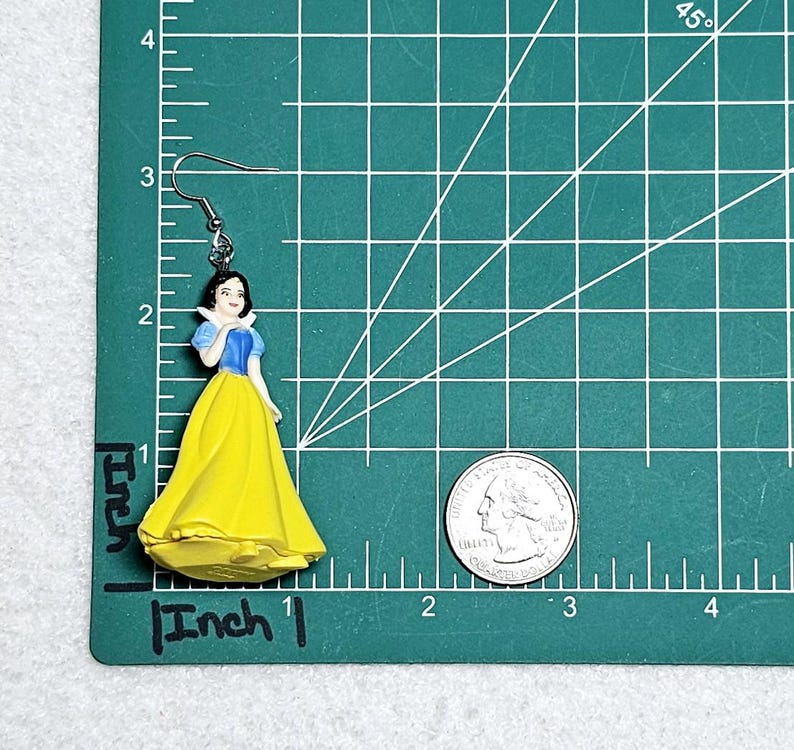 May include: A pair of silver earrings featuring a plastic figurine of Snow White in a yellow dress with a blue bodice. The earrings are approximately 1 inch long.