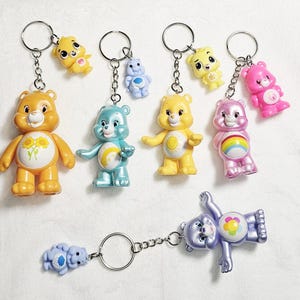 May include: A collection of Care Bears keychains featuring various characters in yellow, blue, pink, and purple. Each keychain includes a metal ring, a chain, and a small plastic bear figure with distinct belly badge symbols like flowers, suns, and rainbows.