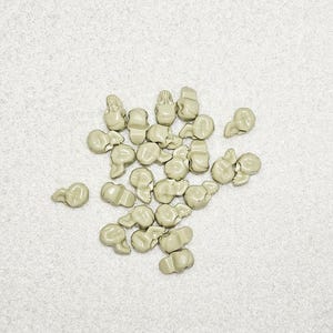 May include: A collection of small, light green-gray ceramic beads scattered on a white textured surface. The beads have a unique, abstract shape, resembling small, stylized figures. The lighting is soft, highlighting the smooth texture of the beads.