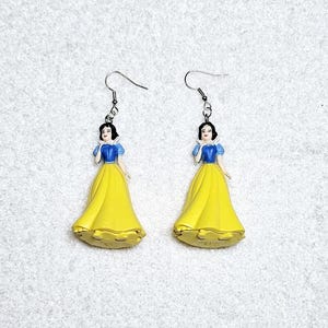 May include: A pair of dangle earrings featuring a plastic figurine of Snow White from Disney's Snow White and the Seven Dwarfs. The figurine is wearing a yellow dress with a blue bodice and a white collar. The earrings have silver hooks.