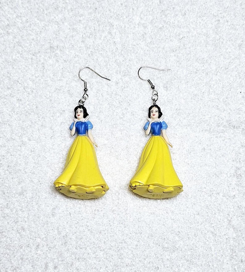 May include: A pair of dangle earrings featuring a plastic figurine of Snow White from Disney's Snow White and the Seven Dwarfs. The figurine is wearing a yellow dress with a blue bodice and a white collar. The earrings have silver hooks.