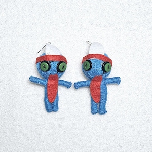 May include: A pair of handmade blue yarn doll earrings. Each doll has a red headband and scarf, green button eyes, and a white hat. The earrings are designed to look like small figures with outstretched arms.