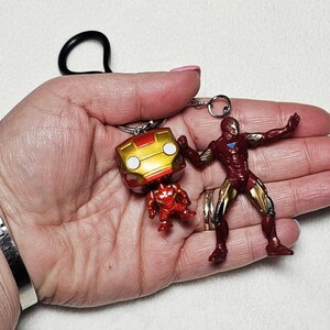 May include: A keychain featuring two Iron Man figures attached to a black ring. One figure is a stylized bobblehead with a large gold and red helmet, while the other is a full-body action figure in a dynamic pose with red and gold armor plating.