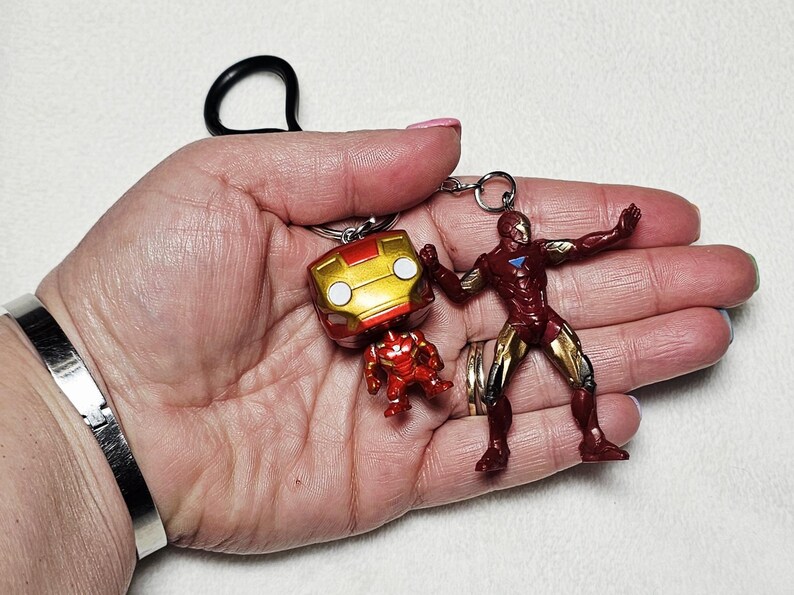 May include: A keychain featuring two Iron Man figures attached to a black ring. One figure is a stylized bobblehead with a large gold and red helmet, while the other is a full-body action figure in a dynamic pose with red and gold armor plating.