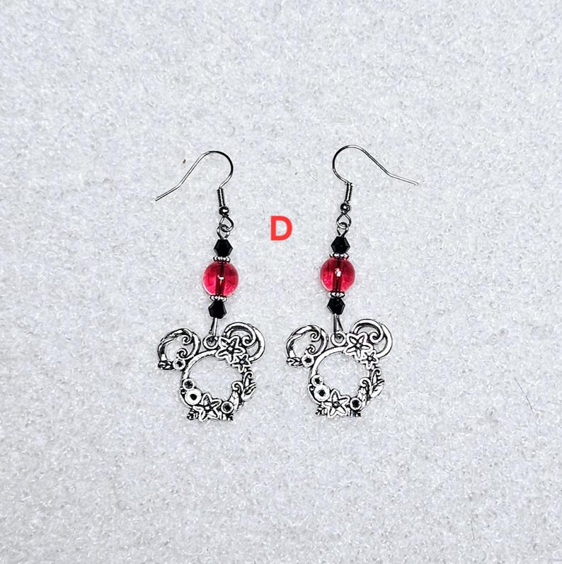 K&ouml;nnte beinhalten: Ein Paar silberne Ohrringe mit einem floralen Design und einer roten Glasperle. Die Ohrringe h&auml;ngen an silbernen Haken.