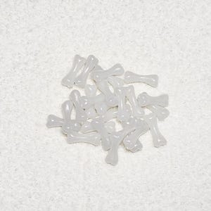 May include: A collection of small, translucent bone-shaped beads. The beads are a pale, off-white color and appear to be made of plastic or resin. They are scattered across a textured, white surface, suggesting a craft or jewelry-making theme.