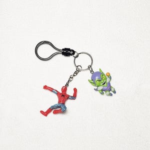 May include: A metal keychain featuring a black lobster clasp and a split ring. Two small character charms are attached to the ring: a red and blue superhero figure in a dynamic pose and a green-skinned villain figure wearing a purple hood.