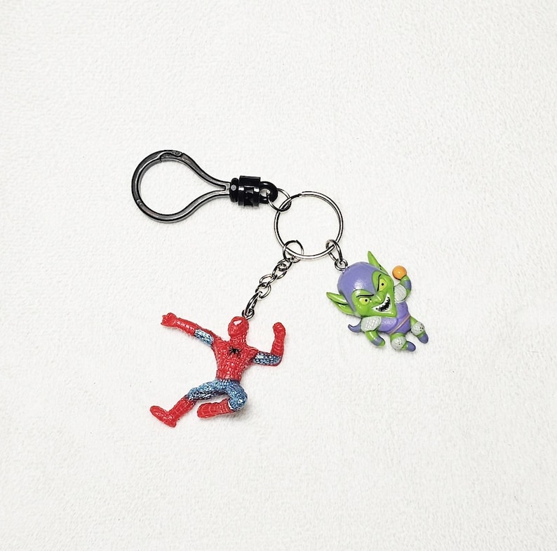 May include: A metal keychain featuring a black lobster clasp and a split ring. Two small character charms are attached to the ring: a red and blue superhero figure in a dynamic pose and a green-skinned villain figure wearing a purple hood.