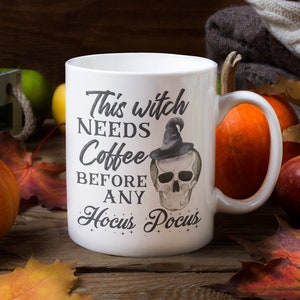 May include: A white ceramic mug with the text "This witch needs coffee before any Hocus Pocus" and a skull illustration. The mug is surrounded by pumpkins, apples, and fall leaves on a wooden surface.
