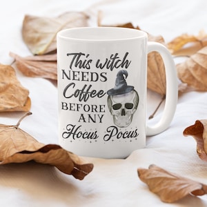 May include: White ceramic mug with the text "This witch NEEDS Coffee BEFORE ANY Hocus Pocus" and a skull wearing a witch hat. The mug is surrounded by dried leaves, creating a fall or Halloween theme.