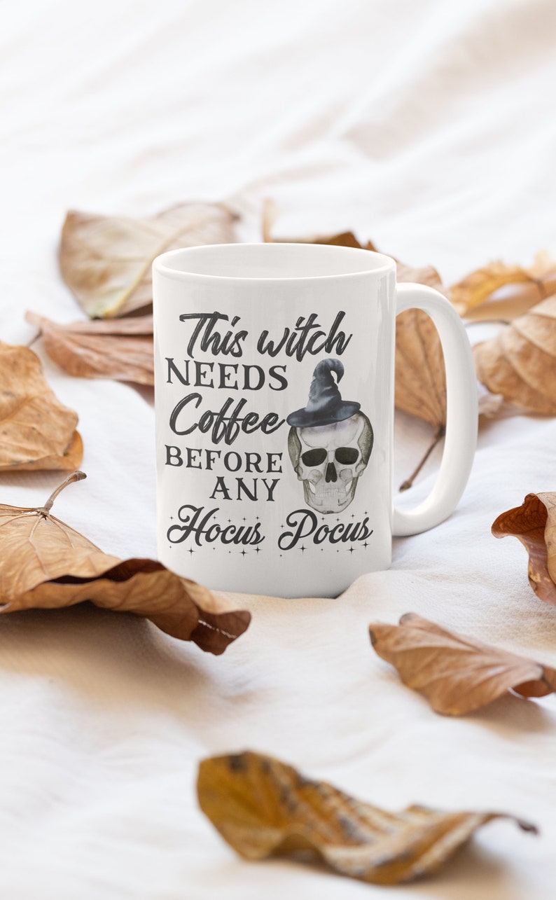 May include: White ceramic mug with the text "This witch NEEDS Coffee BEFORE ANY Hocus Pocus" and a skull wearing a witch hat. The mug is surrounded by dried leaves, creating a fall or Halloween theme.