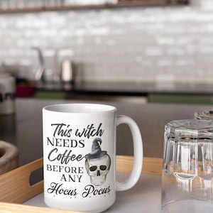 May include: White ceramic mug with the text "This witch needs coffee before any Hocus Pocus" and a skull wearing a witch hat. The mug is on a wooden tray with a glass next to it. Perfect for Halloween or a witch-themed gift.