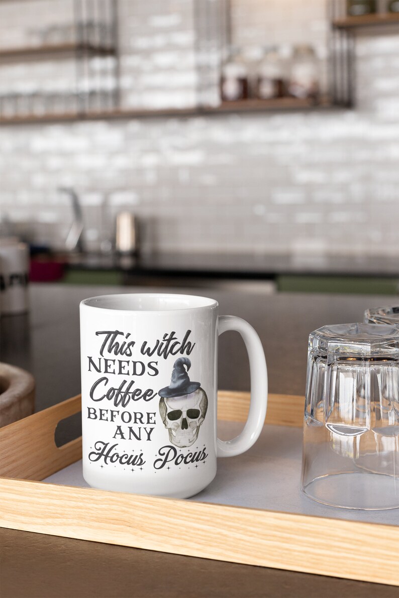 May include: White ceramic mug with the text "This witch needs coffee before any Hocus Pocus" and a skull wearing a witch hat. The mug is on a wooden tray with a glass next to it. Perfect for Halloween or a witch-themed gift.
