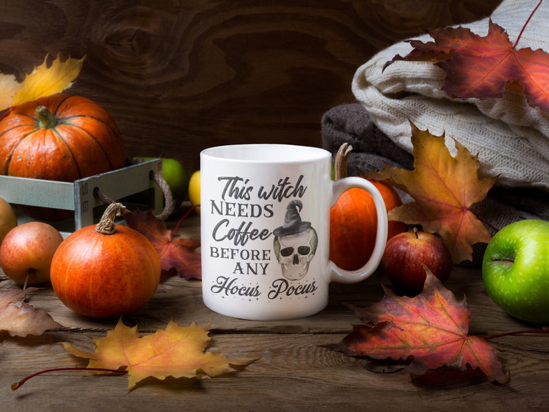 May include: A white ceramic mug with the text "This witch needs coffee before any Hocus Pocus" and a skull illustration. The mug is surrounded by pumpkins, apples, and fall leaves on a wooden surface.