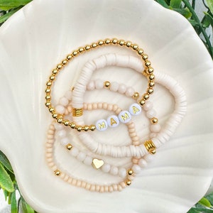 May include: A set of five bracelets with white beads and gold accents. One bracelet features the word "MAMA" in gold letters on white beads. The bracelets are arranged on a white shell-shaped dish.