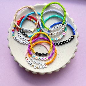 May include: A selection of colourful beaded bracelets, including green, blue, and orange, presented on a white, scalloped plate. Several bracelets display names spelled out with letter beads, such as "ISLA" and "ALLY".
