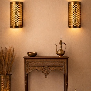 Peut inclure: Deux appliques murales cylindriques dorées avec un motif en losange sont montées sur un mur beige. En dessous, une console en bois avec des sculptures ornementales supporte un bol en laiton, une théière et des pots décoratifs. Un grand vase à motifs avec des plantes séchées est à gauche.