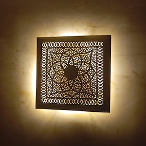 Op de afbeelding: Een vierkante, witte, decoratieve wandlamp met een geometrisch, ingewikkeld, uitgesneden ontwerp. De lamp is verlicht en geeft een warm licht.