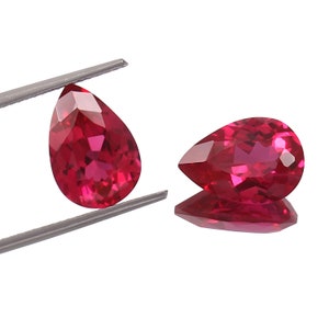 Könnte beinhalten: Zwei birnenförmige rote Edelsteine, jeweils mit einem facettierten Schliff, werden auf einer silbernen Pinzette präsentiert. Die Edelsteine sind von einer leuchtenden, tiefen roten Farbe und haben einen hohen Glanz.