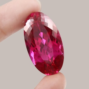 May include: A large, oval-shaped, faceted gemstone with a deep pink hue. The gemstone is held in a hand, showcasing its brilliance and clarity.