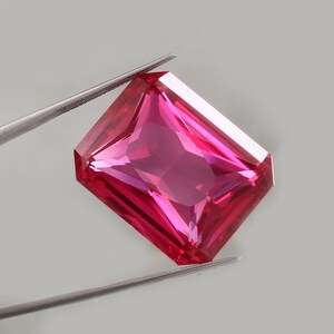 Puede incluir: Una gema rosa brillante, tallada en forma rectangular, sostenida por unas pinzas plateadas. Las facetas de la gema reflejan la luz, creando un efecto brillante. El fondo es gris neutro. La gema es de un rosa intenso.
