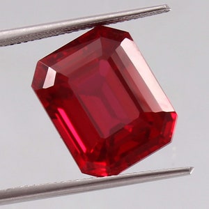 Könnte beinhalten: Ein tief roter Smaragdschliff-Edelstein, der von einer Pinzette gehalten wird. Der Edelstein hat eine leuchtend rote Farbe und eine hohe Klarheit.