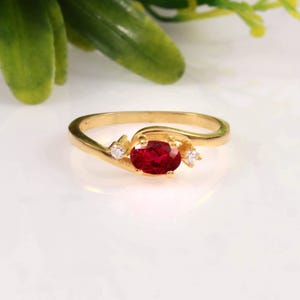 May include: A gold ring featuring a central oval-cut red gemstone held by four prongs, flanked by two small clear round-cut stones on either side. The band has a smooth, polished finish and a bypass design that curves around the centre stones.