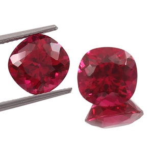 May include: Two cushion-cut red gemstones, each with a brilliant, faceted surface. The gemstones are held by tweezers against a white background.