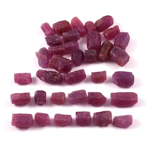 May include: A collection of numerous small, raw ruby crystals in varying shades of deep pink and magenta. The stones are irregularly shaped with rough, textured surfaces and some translucent areas, arranged in loose rows and a cluster on a white surface.