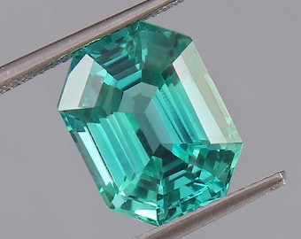 May include: A vibrant green emerald gemstone with a rectangular emerald cut. The gemstone is held in a pair of tweezers.