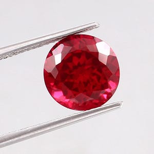 May include: A round, faceted, deep red gemstone is held by a pair of silver tweezers against a white background. The gemstone's facets reflect light, creating a brilliant display of color and sparkle. This image showcases a precious stone.
