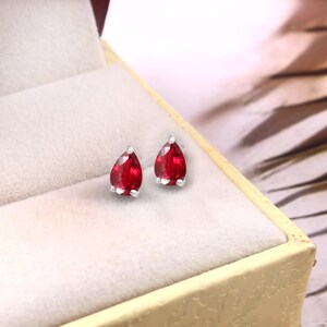 May include: A pair of teardrop-shaped ruby earrings with a silver setting. The vibrant red gemstones are displayed inside a cream-coloured jewellery box, creating a striking contrast. The earrings are a classic jewellery item.