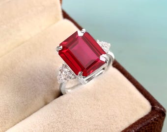May include: A silver ring with a large, rectangular, deep red gemstone as the focal point. The ring features smaller, clear accent stones on either side of the main stone. The ring is presented in a brown jewelry box.