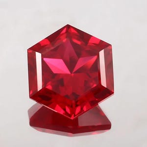 May include: A faceted, hexagonal-cut ruby gemstone. The gem is a vibrant red color with internal facets creating a starburst effect. The gem is displayed on a reflective surface, showcasing its brilliance.