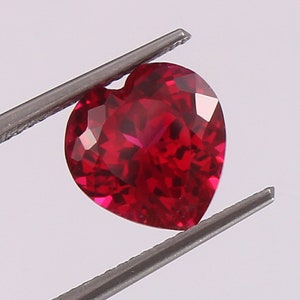 May include: A heart-shaped ruby gemstone with a deep red color. The gemstone is held in place by a pair of tweezers.