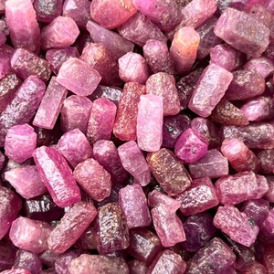 May include: A collection of raw, unpolished ruby gemstones in various shades of pink and deep red. The stones feature irregular, blocky shapes with textured, crystalline surfaces and varying degrees of translucency, packed closely together.