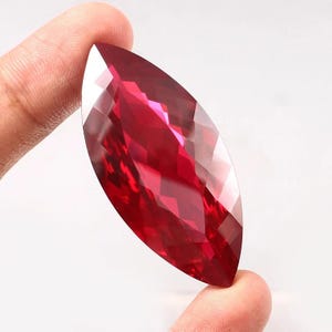 Puede incluir: Una gema facetada con corte marquesa de color rojo intenso. La gema se sostiene entre dos dedos, mostrando su forma alargada y múltiples facetas que reflejan la luz. La piedra preciosa parece ser un rubí o una piedra preciosa similar.