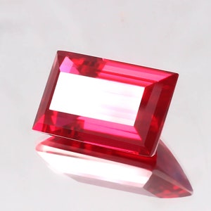 May include: A deep red gemstone with a rectangular emerald cut. The gemstone is faceted and has a high polish.