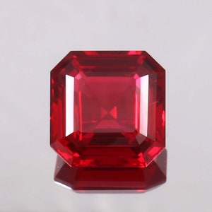 Könnte beinhalten: Ein tiefroter Edelstein mit einem quadratischen Smaragdschliff. Der Edelstein ist facettiert und hochglanzpoliert.