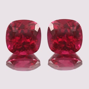 May include: Two vibrant red cushion-cut gemstones with a high polish. The gemstones are set against a white background and are reflected in the surface below.