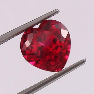 May include: A heart-shaped, faceted ruby gemstone held by tweezers. The ruby is a deep red color and has many reflective surfaces. The tweezers are silver and the background is a light gray.
