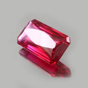 Puede incluir: Una gema rectangular facetada con un vibrante tono rosa intenso. La gema es muy reflectante, con bordes afilados y una superficie pulida. El reflejo de la piedra es visible debajo, creando un efecto simétrico. La gema está sobre un fondo neutro.