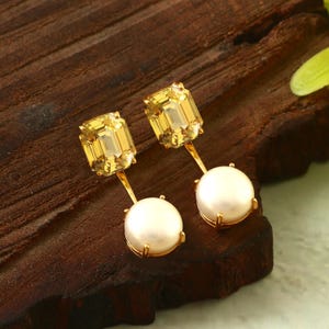 May include: A pair of gold-toned earrings. Each earring features a large, rectangular, yellow gemstone above a round, white pearl. The earrings are displayed on a dark wooden surface.