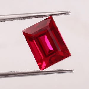 May include: A rectangular-cut ruby gemstone held by tweezers. The deep red gemstone has a faceted surface, reflecting light. The ruby's shape is an emerald cut, showcasing its clarity and brilliance. The tweezers are silver.