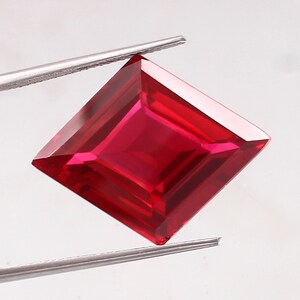 May include: A deep red square-shaped gemstone with a faceted cut. The gemstone is held by a pair of tweezers.