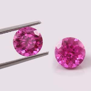 May include: Two round, pink gemstones with a faceted cut. The gemstones are held by tweezers.