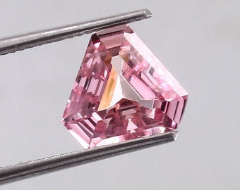 May include: A pink triangular gemstone with a faceted cut, held by tweezers against a white background.