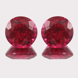 May include: Two round, faceted ruby gemstones with a deep red color. The gemstones are set against a white background.